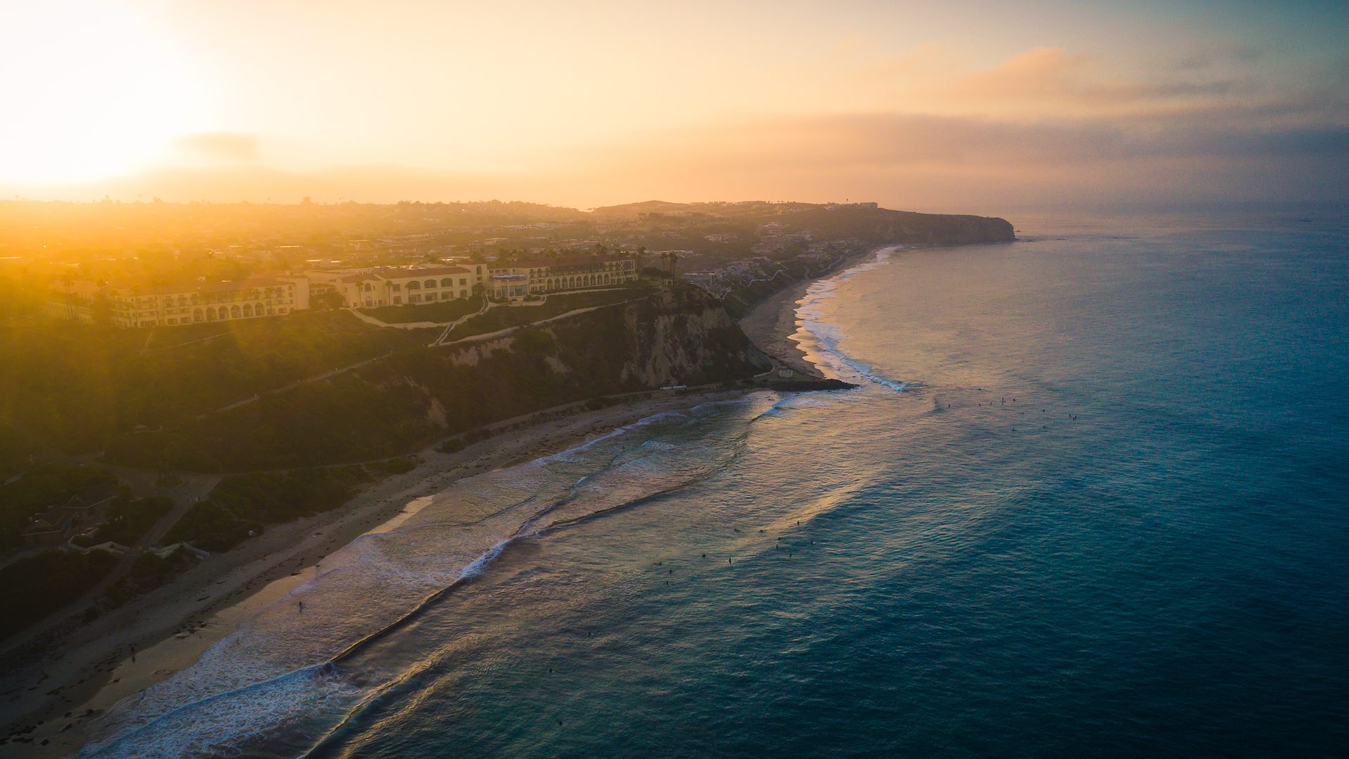 Golden-hour aerial of the Dana Point coastline: cliffs, a crescent beach with white surf, and bluff-top buildings under a hazy sun for the home page of  Three Points Wealth Planners, based in Dana Point